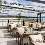 Wicker chairs with cushions line a rooftop terrace, under a pergola adorned with lights and greenery. The backdrop features a scenic view of distant trees and water.