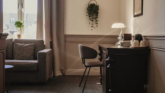 Ett mysigt hotellrum med en bekväm soffa vid ett solbelyst fönster och en funktionell skrivbordsplats med lampa och förfriskningar.
