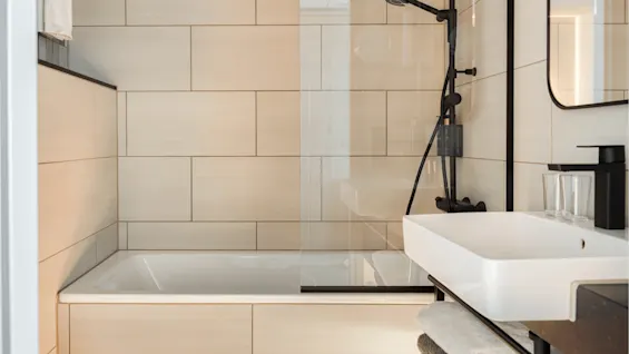 Bathtub surrounded by beige tiles, fitted with a black shower head, adjacent to a white sink with a black faucet. A towel hangs on a rack above the tub.