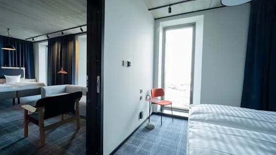Modern hotel room with two beds, a sofa, and a large window offering natural light. Blue and white decor.