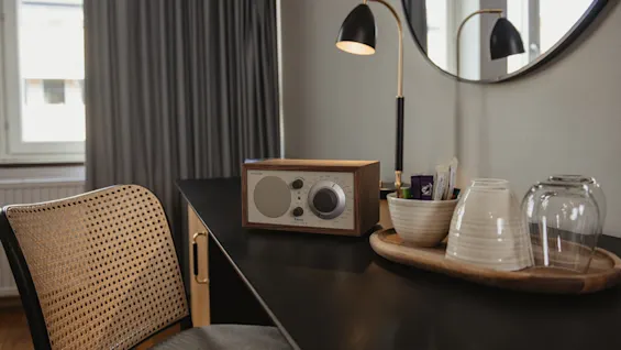 En radio står på et svart skrivebord ved siden av en flettet stol i et mykt opplyst rom. I nærheten står et brett med kopper og pakker under en stilren lampe og rund speil.