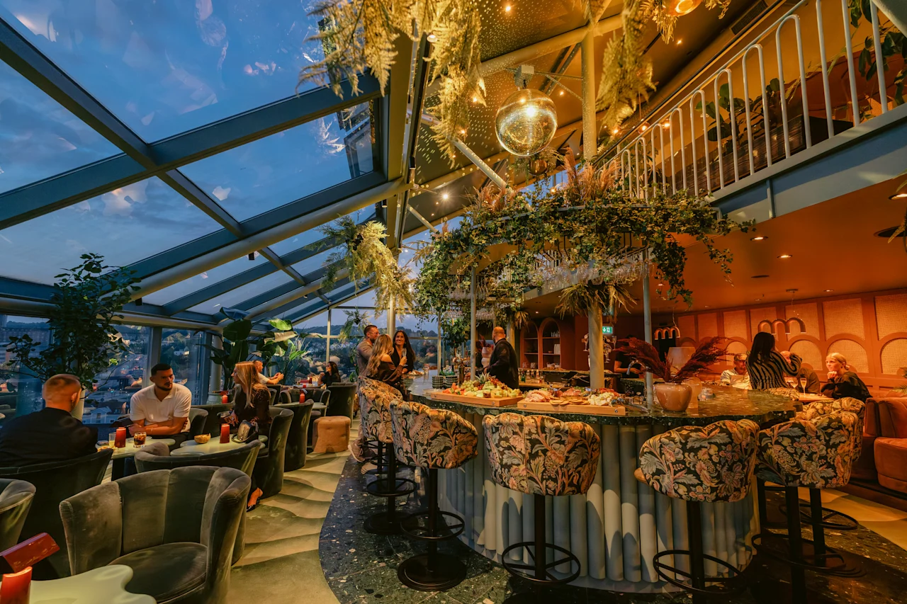 Ihmiset nauttivat juomia ja ruokaa vilkkaalla kattobaarissa, jossa on lasikatto, rehevää kasvillisuutta ja näkymä iltataivaalle.