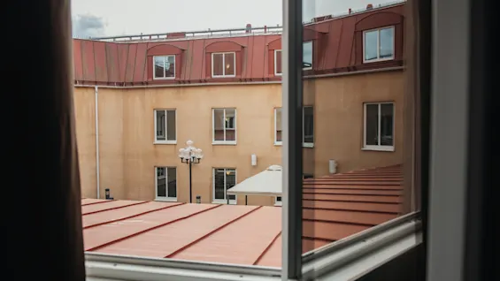En innergård syns genom ett öppet fönster med en beige byggnad och röda tak. Fönster kantar byggnaden och en gatlykta står på den stenlagda gården.