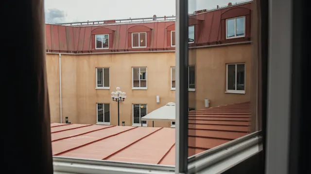 A courtyard view through an open window, showing a beige building with red roofs. Windows line the building, and a streetlamp stands on the courtyard's paved area.