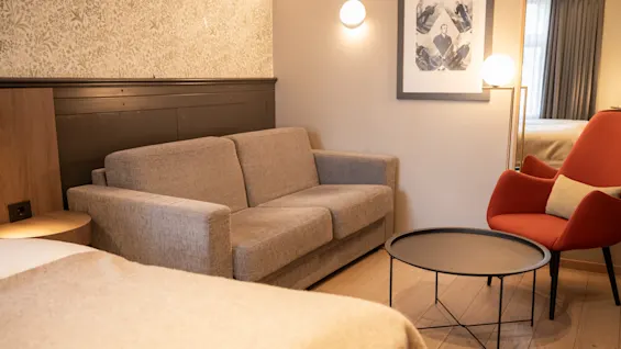 En grå soffa står bredvid ett modernt runt bord i ett mysigt rum, tillsammans med en röd fåtölj och väggkonst. Varm belysning skapar en inbjudande atmosfär.