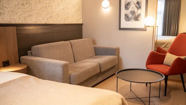 En grå sofa står ved siden av et moderne rundt bord i et koselig rom, sammen med en rød lenestol og veggkunst. Varm belysning skaper en innbydende atmosfære.