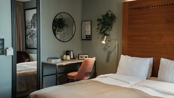 Et hyggeligt hotelværelse med en seng, et skrivebord med en elkedel og radio, og et spejl, der reflekterer rummets indbydende atmosfære.