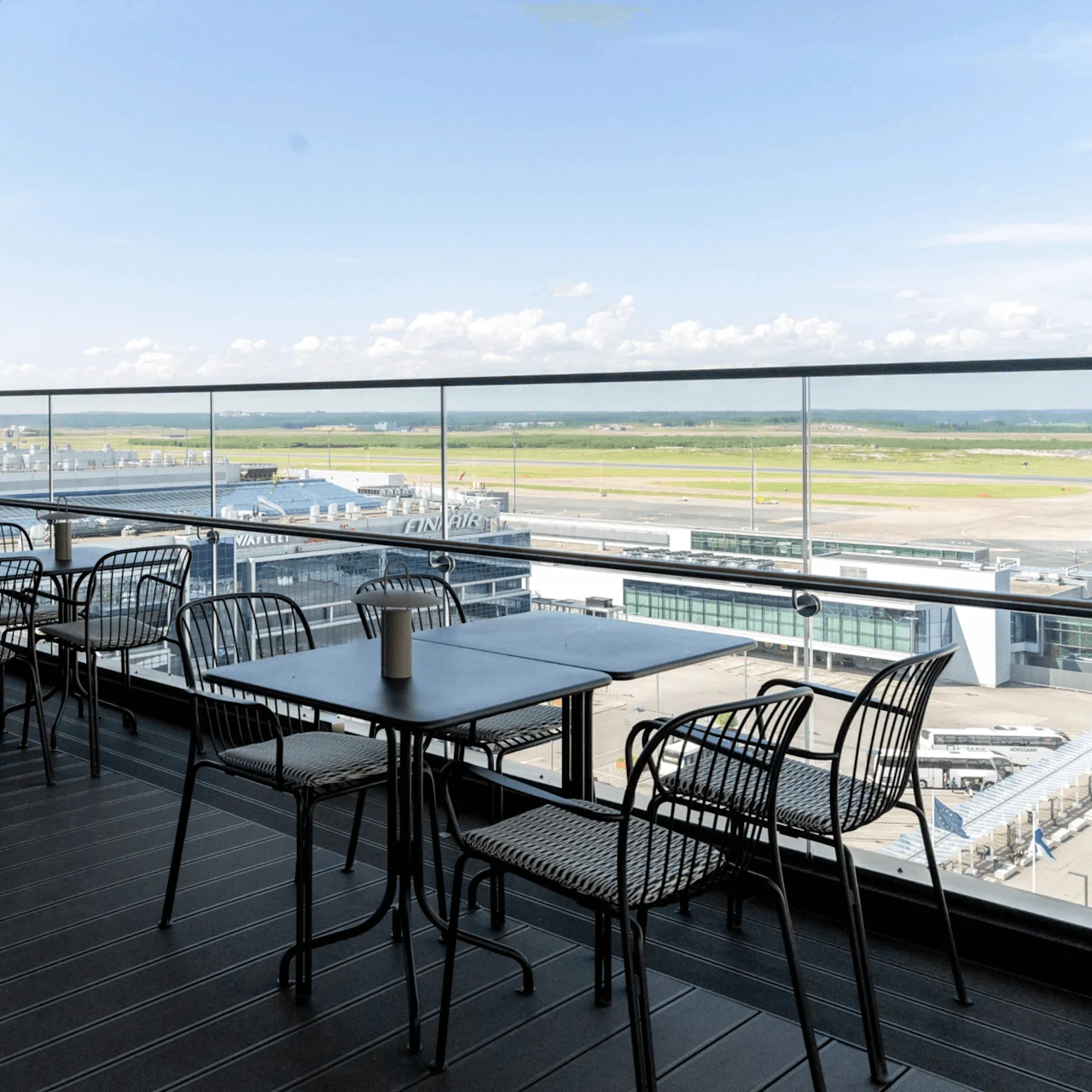 Tables and chairs are set on a rooftop terrace, overlooking an airport runway. A glass railing offers a clear view. Visible building shows text: "AVIA PILOT FINAVIA."