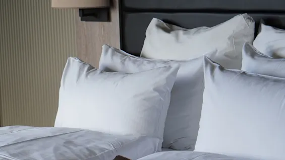 A neatly made bed with fluffed white pillows and a brown throw blanket resting on it. The headboard and a wall lamp provide a cozy, modern bedroom setting.