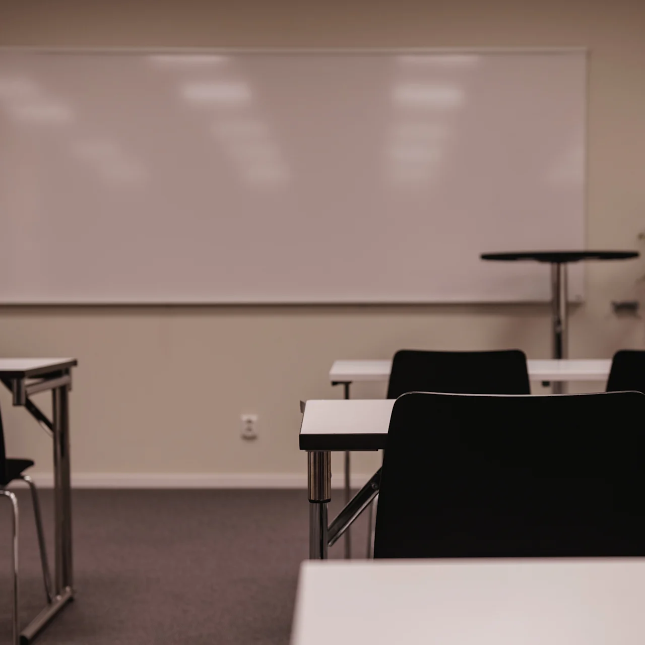 Tables and chairs are arranged in rows facing a whiteboard in a classroom setting. The room is empty, with neutral-colored walls and carpeted floor.