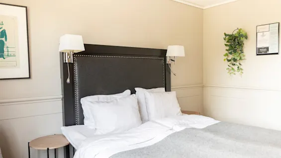 Ett mysigt sovrum med en prydligt bäddad säng, med en mörk sänggavel, vita sängkläder och vägglampor, vilket antyder komfort och avkoppling.