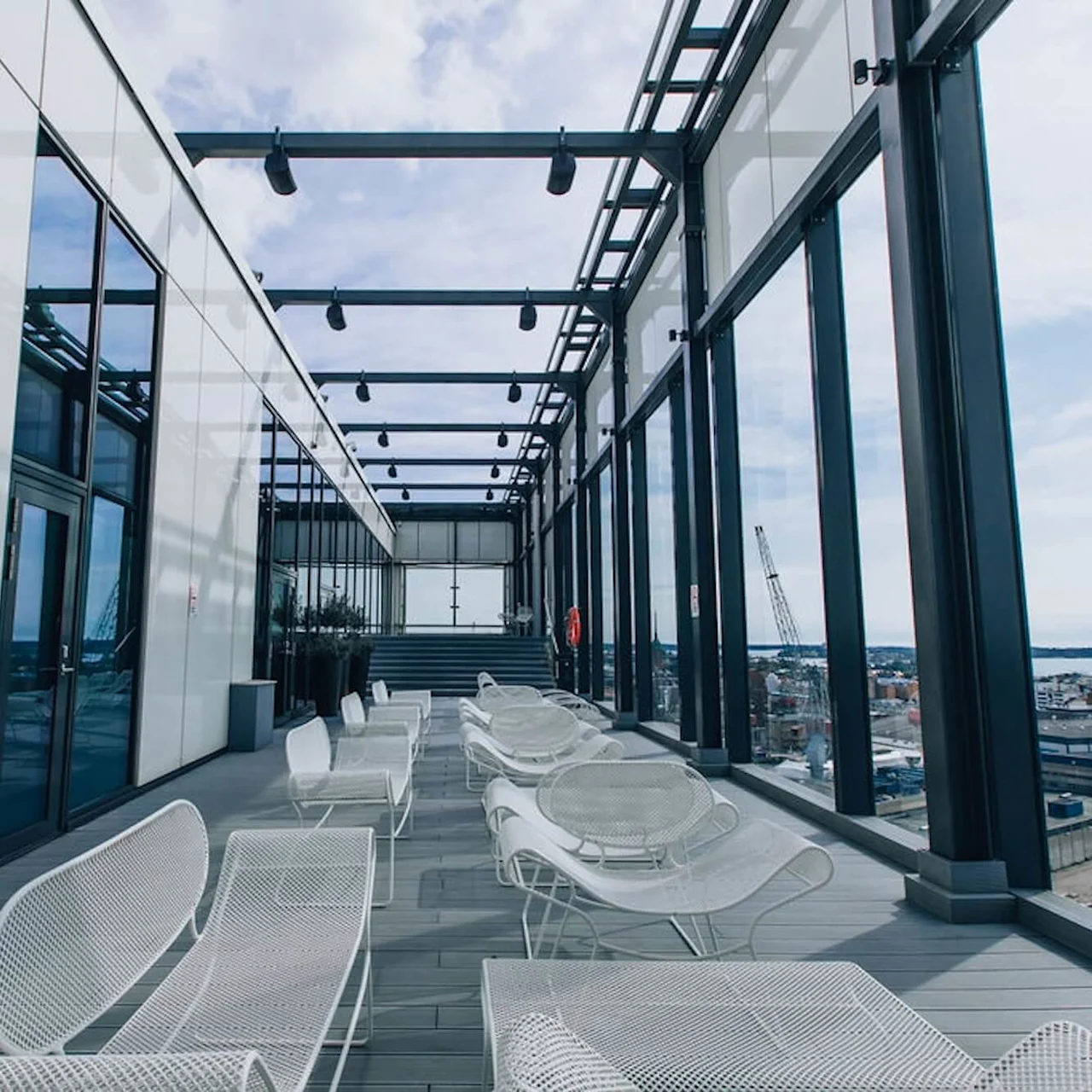 Hvide metalstole er arrangeret på en tagterrasse med udsigt over en industrihavn, omgivet af glasvægge og bjælker under en delvist overskyet himmel.