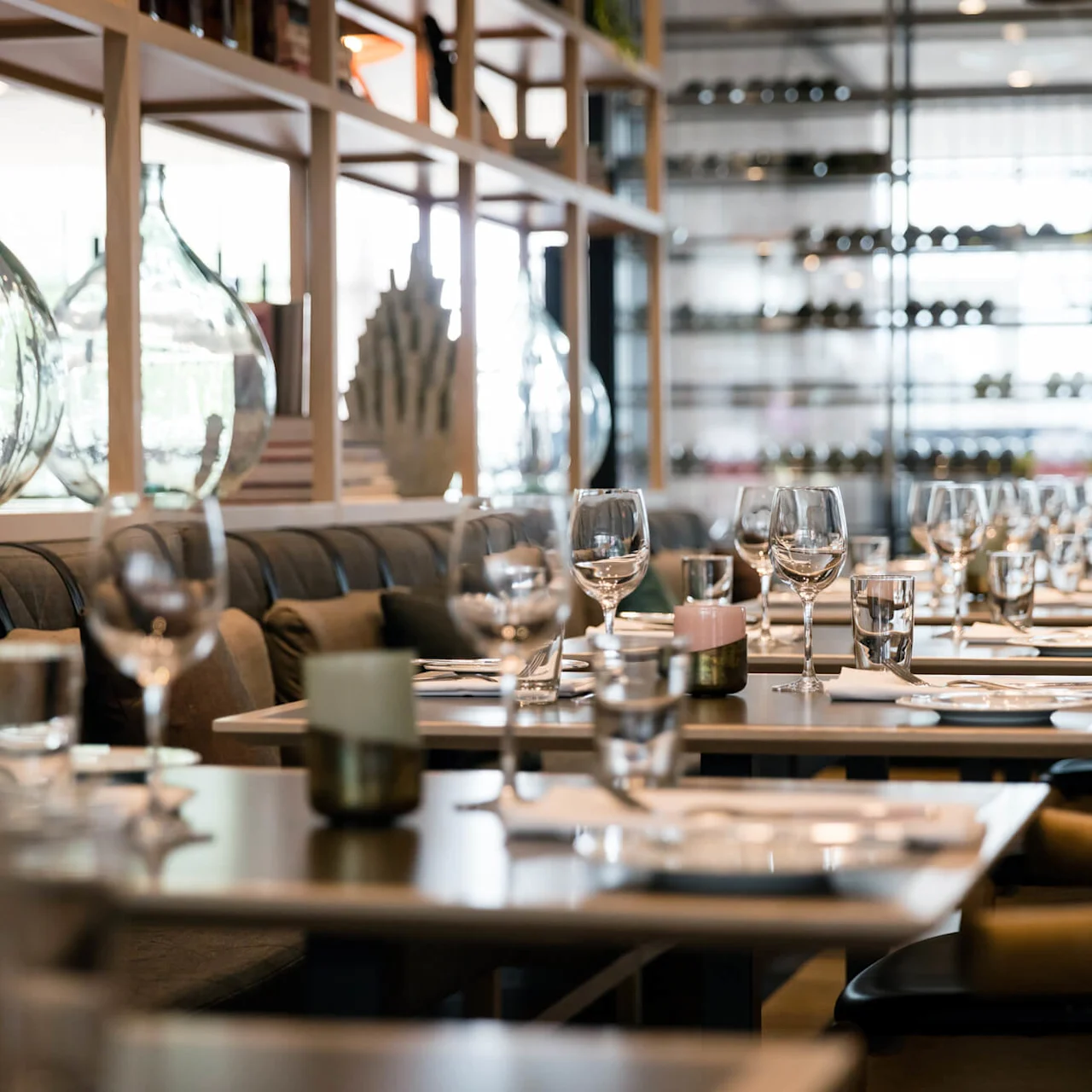 Spisebord, dekket med glass og bestikk, er arrangert i en sofistikert restaurant. Hyller med store glassflasker og en vinutstilling skaper en elegant atmosfære.