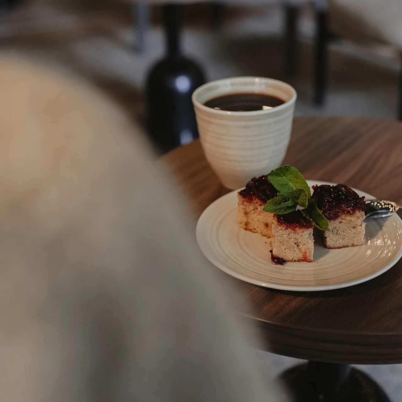 A cup of black coffee and a plate with berry-topped cake slices and mint are on a wooden table in a cozy cafe setting.
