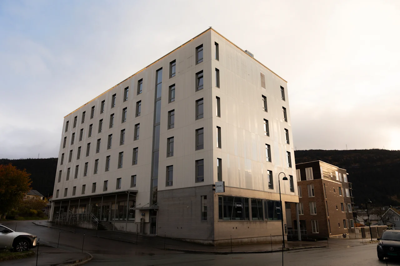 En moderne fleretagers bygning med mange vinduer og skilt Helma, står ved en gade med biler. Bakker i baggrunden.