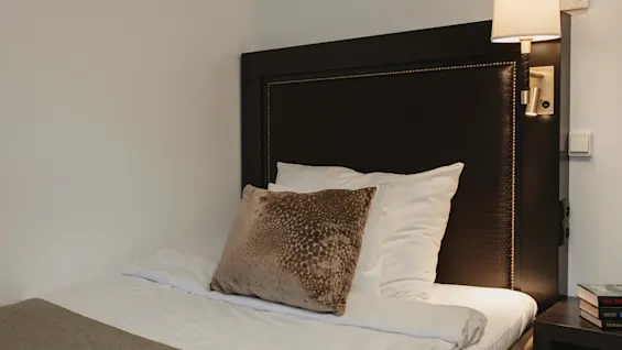 Viihtyisä hotellisänky tummalla sängynpäädyllä, valkoisilla liinavaatteilla ja koristetyynyllä, valaistuna lampulla.