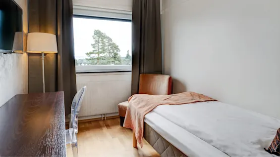 En pent redd seng står mot en vegg, ved siden av et vindu med trær synlige utenfor. Et skrivebord, lampe og stol fullfører det enkle, koselige rommet.