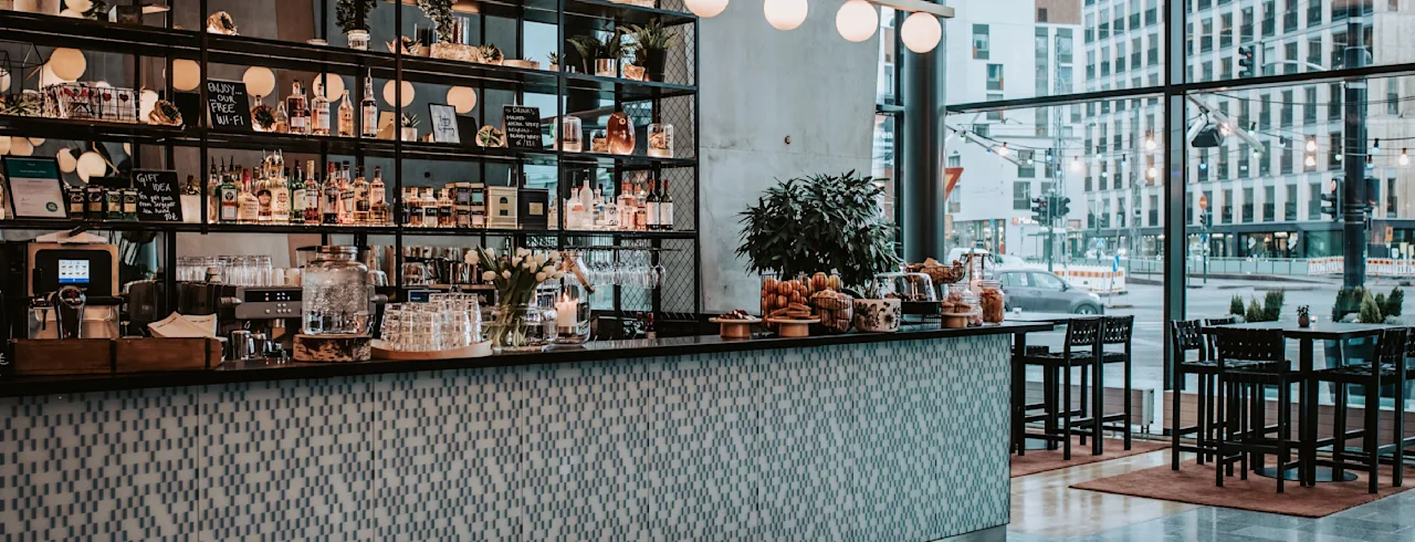 En modern bar har en kaklad disk med hängande lampor, fyllda hyllor och växter; bredvid finns stora fönster som vetter mot en stadsgata. Text på en matta lyder: "SIT BACK, RELAX AND ENJOY."