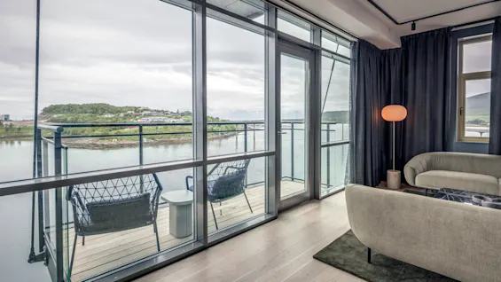 En moderne stue har en hyggelig sofa og en glødende gulvlampe. Glasdøre åbner til en altan med stole, der vender ud mod en rolig vandkant og fjerne grønne områder.