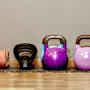 A black kettlebell labeled "4 KG" sits between two larger kettlebells. An orange foam roller lies parallel nearby, all on a wooden floor against a beige wall.
