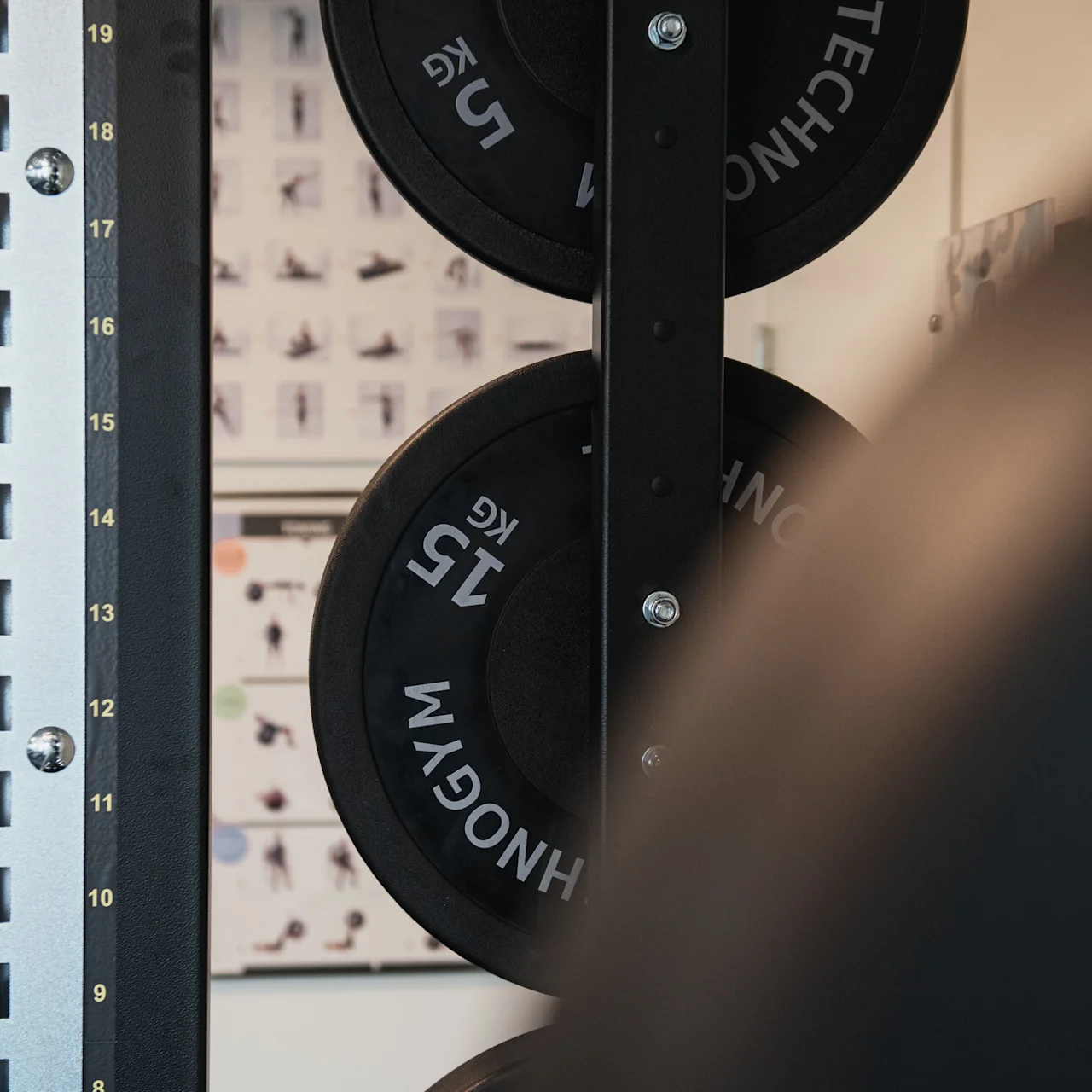 Weight plates resting on a gym rack, with numbers 15 KG visible, against a backdrop of exercise posters on a wall.
