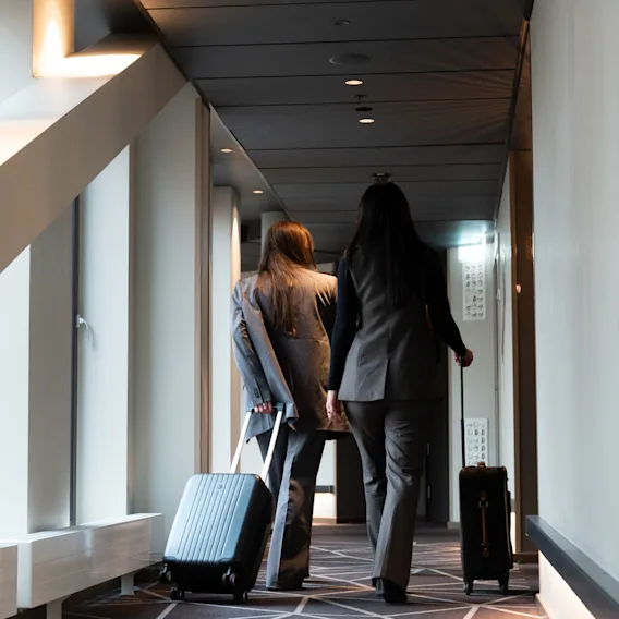 Två kvinnor i kostym går längs en hotellkorridor med geometrisk matta och drar rullväskor efter sig.