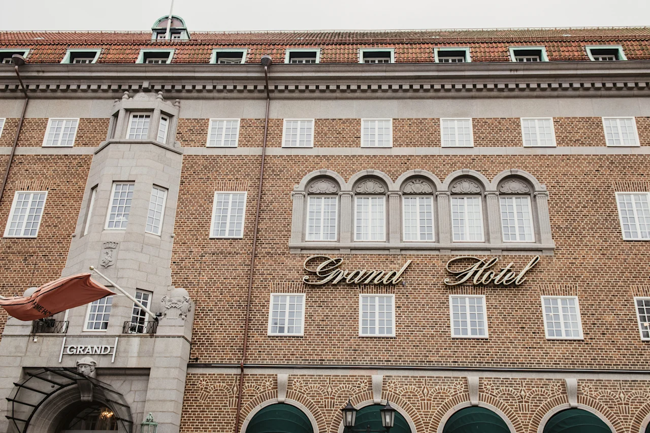 Den ståtliga tegelfasaden på ett hotell, med många fönster och en framträdande skylt där det står 'Grand Hotel'. Entrén säger 'GRAND'.