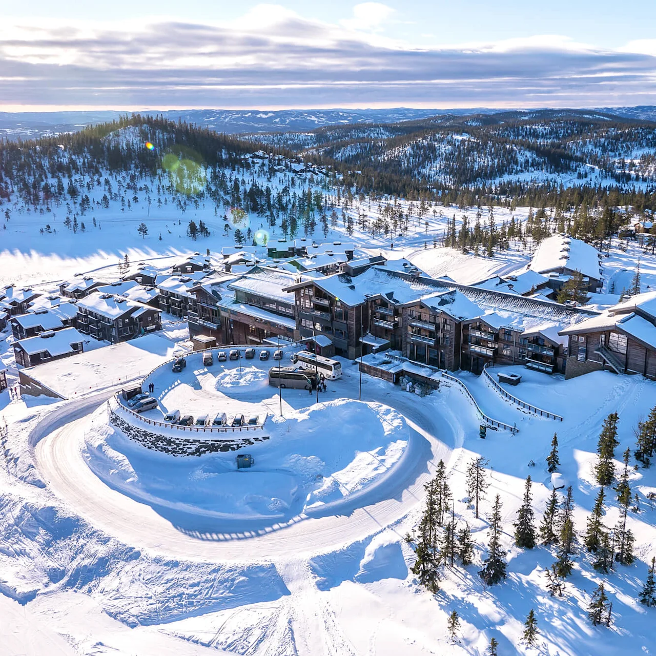 A large, snow-covered resort complex is nestled in a mountainous landscape with pine trees, surrounded by winding snowy paths and parked vehicles. 