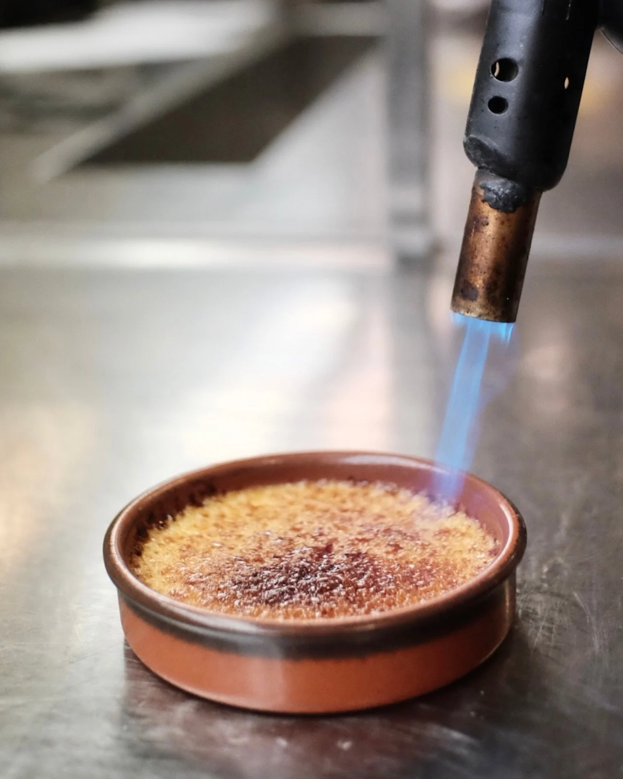 A kitchen torch caramelizes sugar atop round crème brûlée in a brown dish. The dish sits on a metal counter in a kitchen setting.