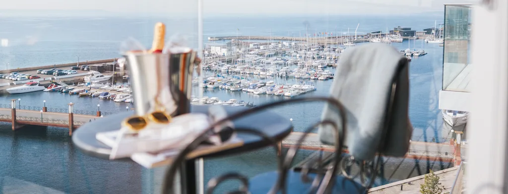 En balkongutsikt over en travel marina med champagne, solbriller og en avis på et bord, som inviterer til avslapning.