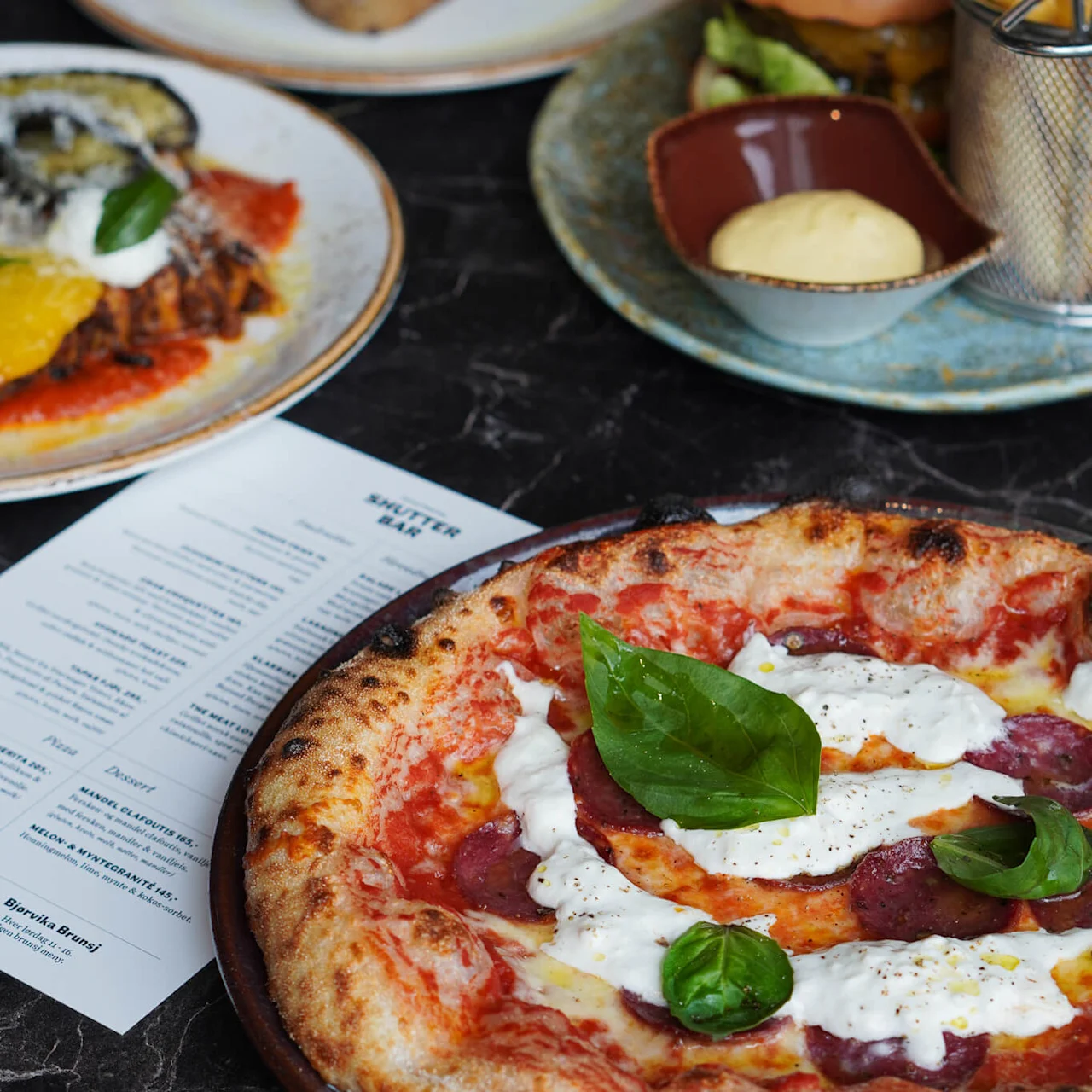 A pizza with salami, white cheese, and basil sits on a black marble table with a menu. Nearby are a burger, fries, and avocado toast.