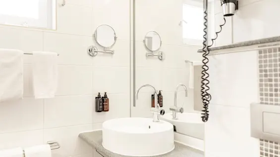 Ett välutrustat hotellbadrum med modernt handfat, stor spegel, väggmonterad hårtork och toalettartiklar.