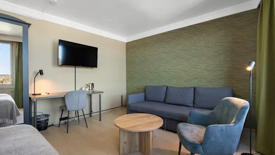 Et moderne hotellrom med en grå sofa, en blå lenestol, et rundt trebord og et skrivebord med en TV over.