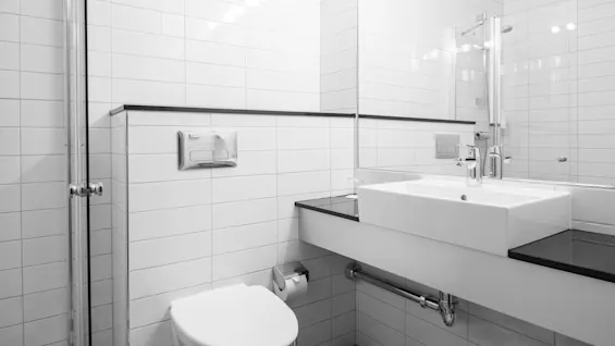 Ett badrum med vita kaklade väggar har en vägghängd toalett, ett fyrkantigt handfat med kran på en svart bänk och en duschvägg av glas. En stor spegel reflekterar rummet.
