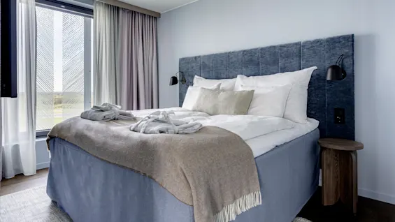Bed adorned with beige throw and white pillows, sits against a blue tufted headboard. Natural light filters through large window with gray curtains, beside a wooden nightstand.