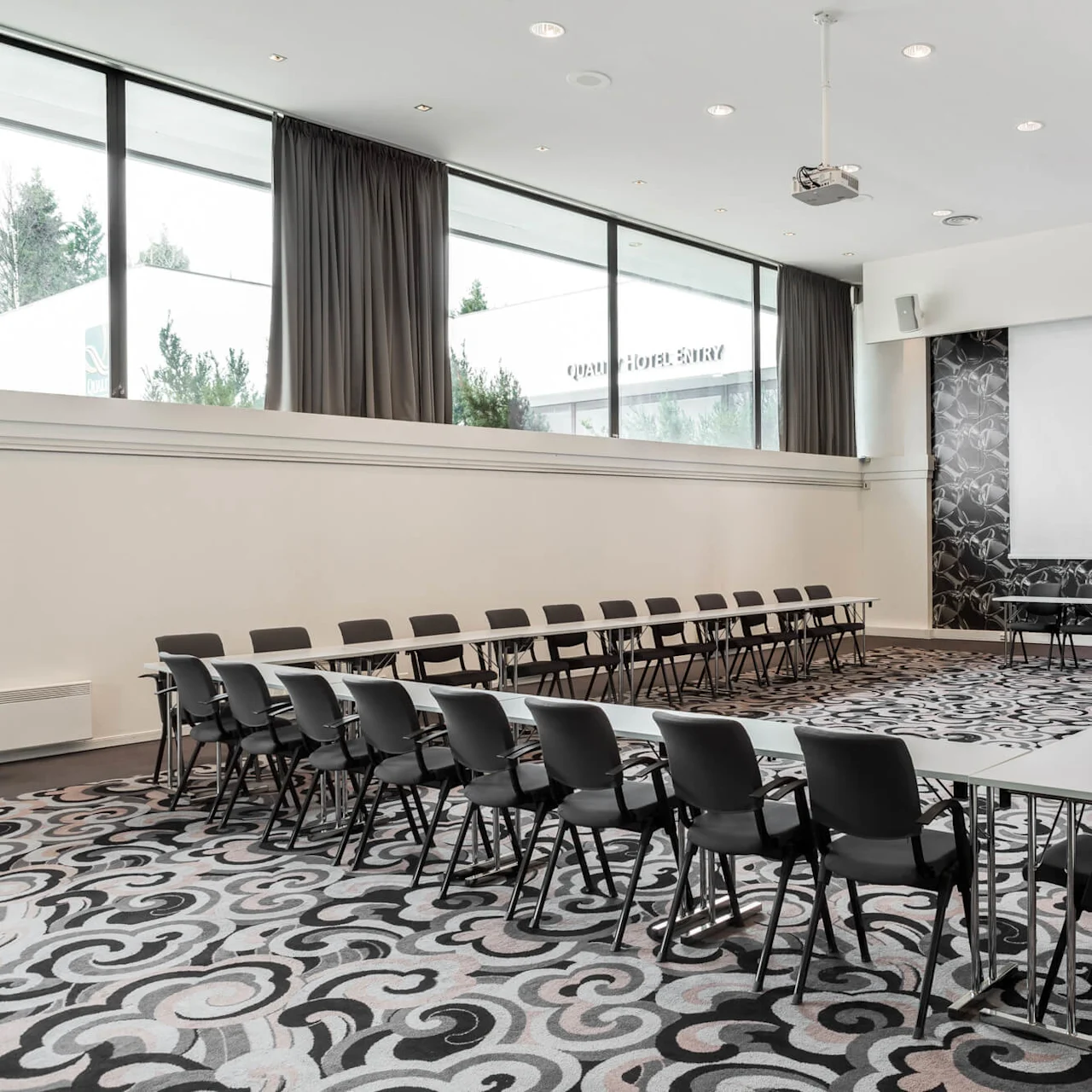 Rows of black chairs surround rectangular tables in a spacious, carpeted conference room with a large screen and patterned accent wall. Windows reveal trees outside.