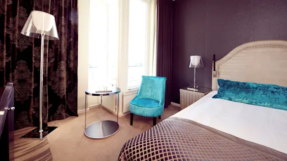 Elegant bedroom with a neatly made bed, teal armchair, floor lamp, and side table, positioned against dark patterned curtains and textured walls.