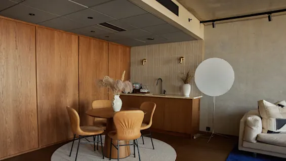 Ett runt träbord med fyra stolar står på en rund matta bredvid en pentry. En stor, vit cirkelformad lampa står nära en beige soffa.