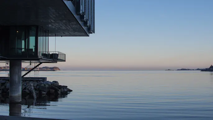Modern building overhanging calm sea at dusk, pilings and rocks visible, tranquil ocean horizon.