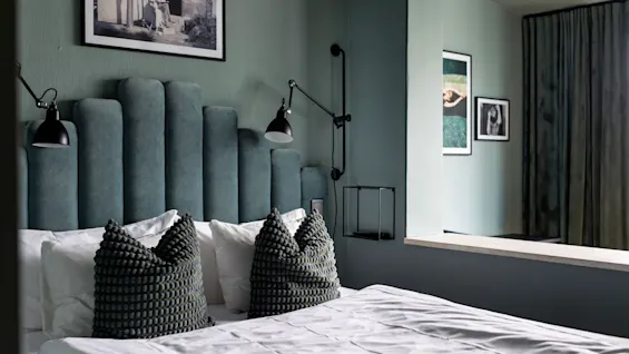 Et moderne hotellrom med en seng med en unik grønn polstret sengegavl, hvitt sengetøy og kunstneriske innrammede trykk på veggene.