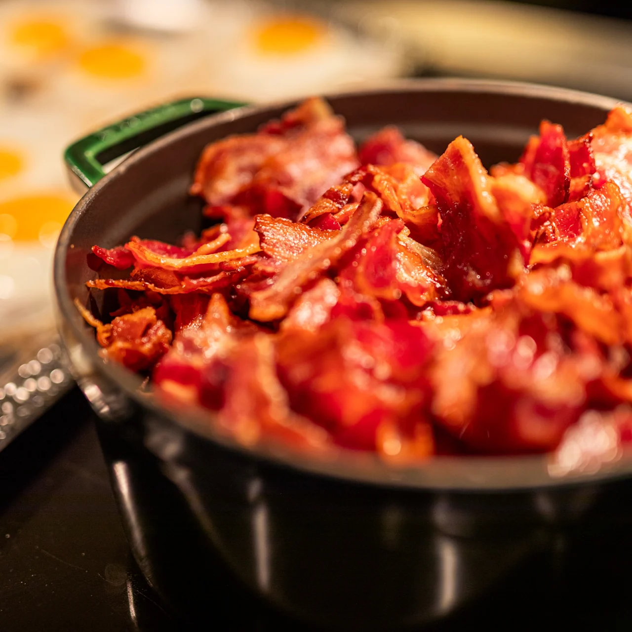 Sprø bacon er stablet i en gryde med grøn kant, placeret på en køkkenbord. I baggrunden er flere spejlæg pænt arrangeret på en bakke.