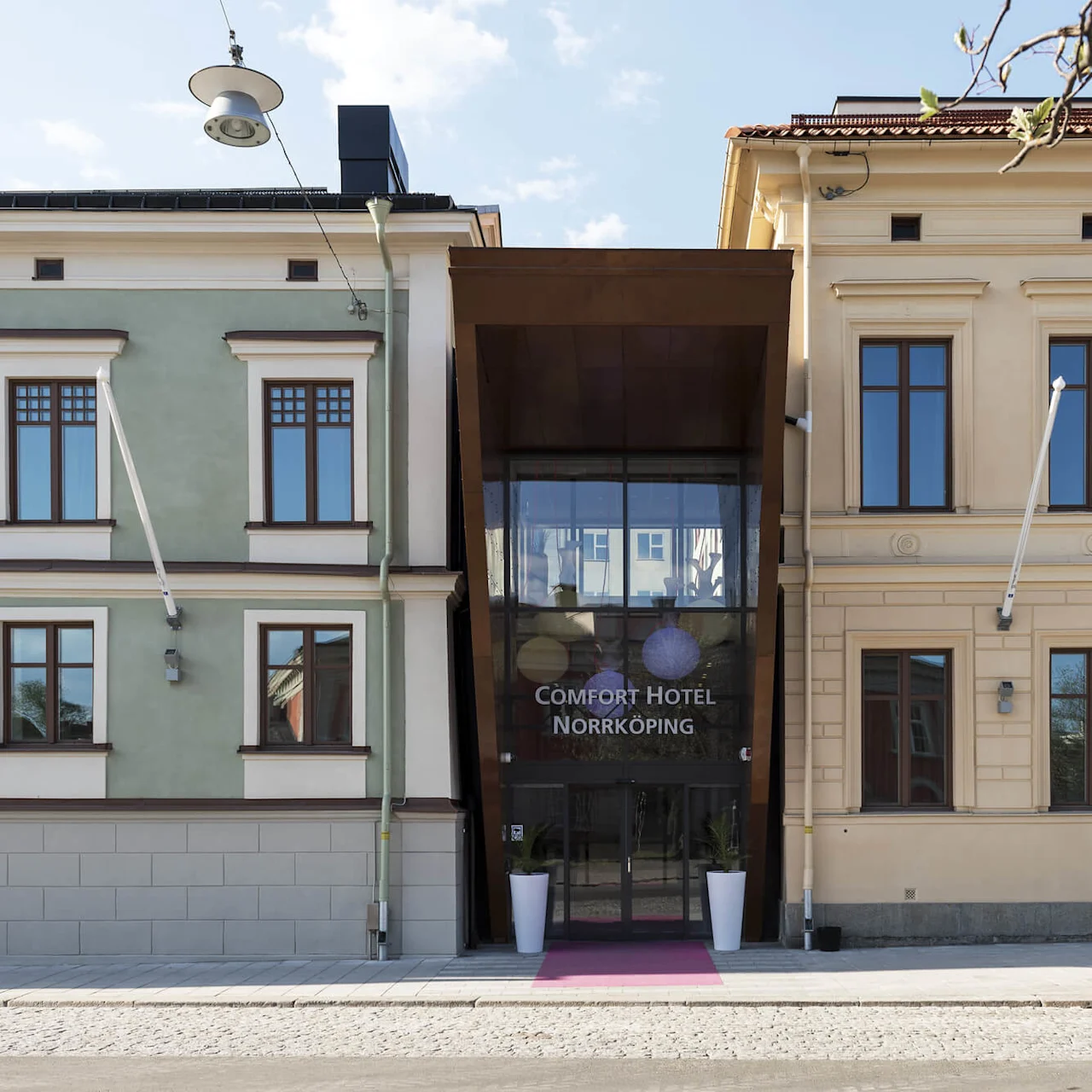 Inngang til Comfort Hotel med en rosa løper og to plantekasser, i en urban setting med historiske bygninger.