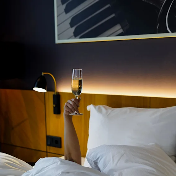 En person i sängen höjer ett glas champagne, med en inramad pianotangentbild ovanför.