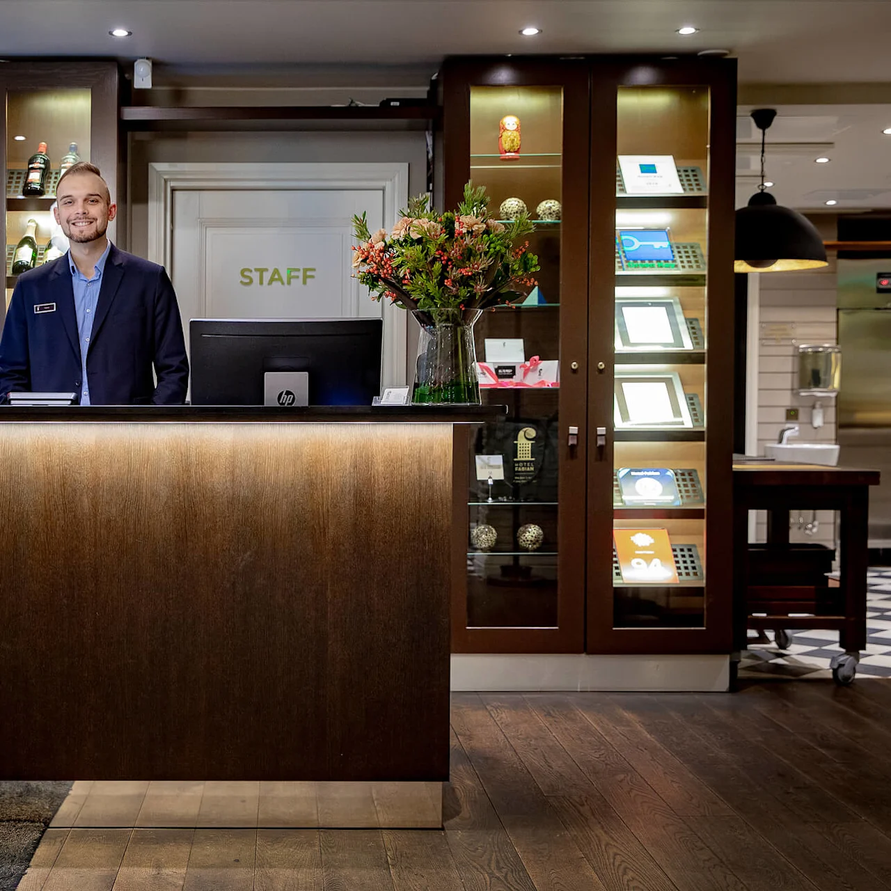 En smilende resepsjonist står ved en trepult, dekorert med blomster, i en hotellobby. Bak er en utstillingshylle med glass og priser. En dør merket "STAFF" er synlig.