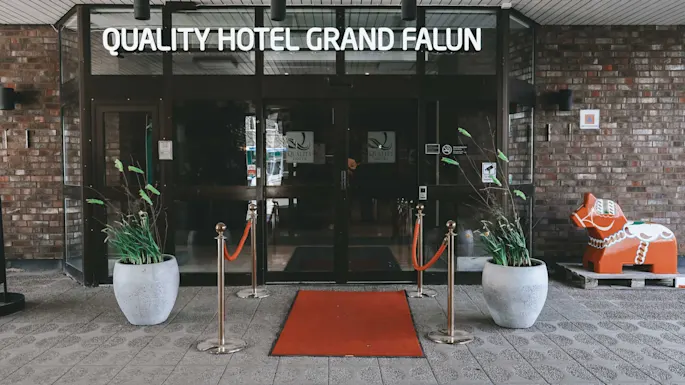 Glassdør med rød løper som leder til "QUALITY HOTEL GRAND FALUN." Flankert av potteplanter; til høyre, en fargerik Dalaheststatue mot en murvegg.