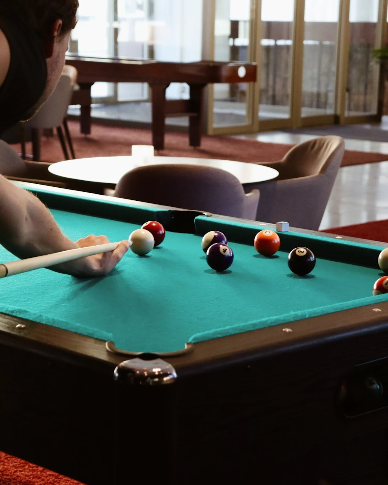 A person holds a pool cue, aiming at billiard balls on a green felt table in a lounge with chairs, a round table, and glass doors in the background.