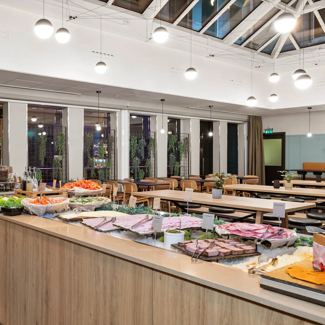 Buffet, jossa on erilaisia ruokia esillä modernissa sisustuksessa, jossa on puisia pöytiä, suuria ikkunoita ja kattolamppuja.