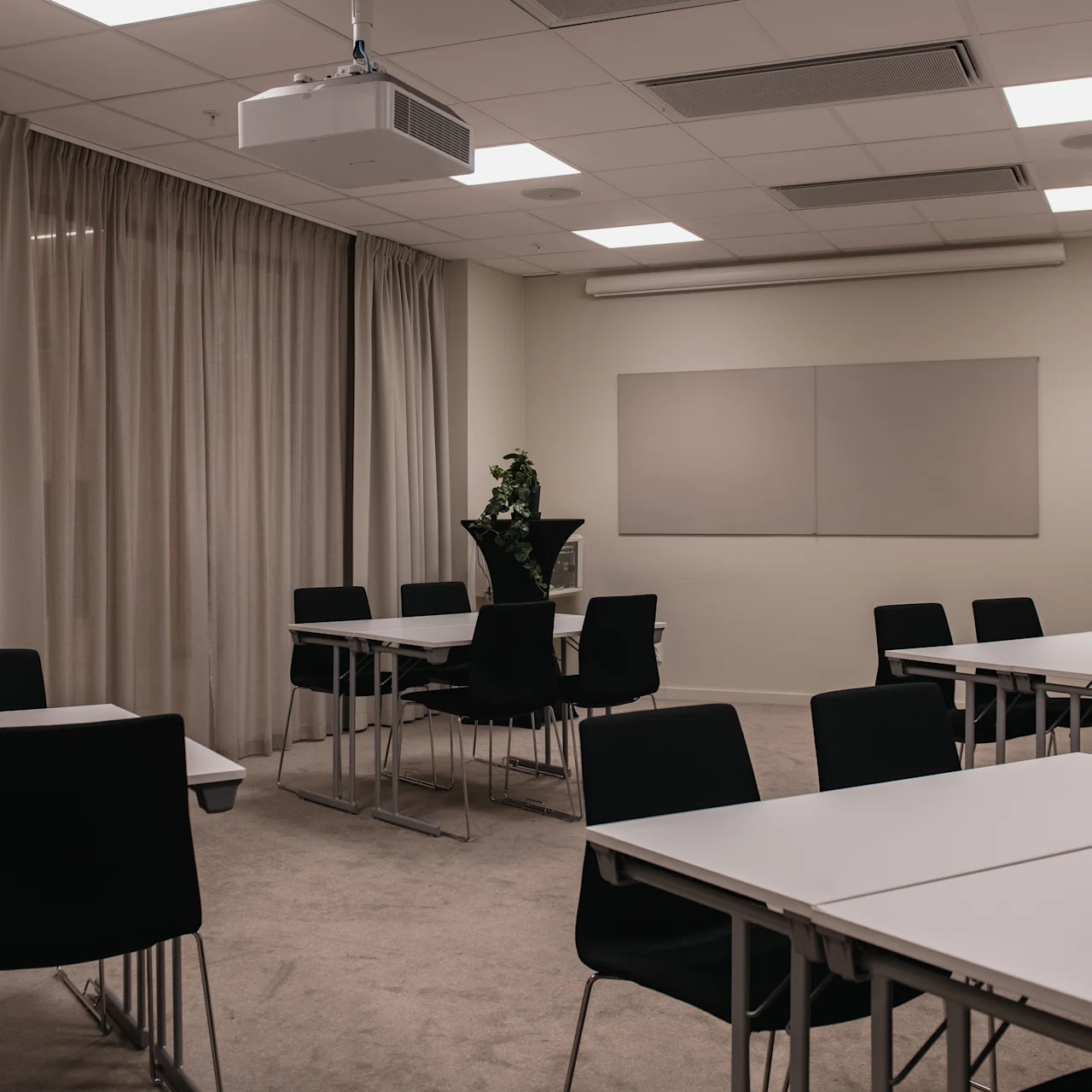 Conference room has black chairs around rectangular white tables. A projector hangs from the ceiling. Beige curtains cover windows; walls are plain. Ceiling lights illuminate the carpeted space.