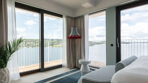 A bedroom with a bed, decorative lamp, and potted plant overlooks a scenic view through large glass doors, opening to a balcony with a lake and distant hills.