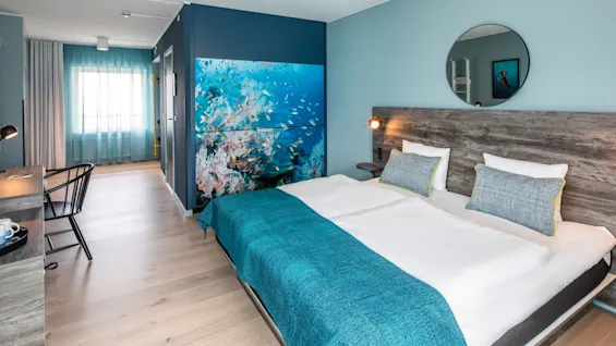 A neatly made bed with turquoise bedding rests under a circular mirror. Wall art depicting an underwater scene adorns a light blue hotel room, complemented by a desk and chair.